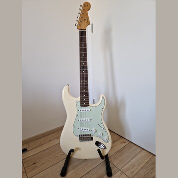 Bílá elektrická kytara s jasně viditelným logem Fender na hlavě. Nástroj má hnědý hmatník, světle zelený pickguard a tři snímače. Kytara je umístěna…