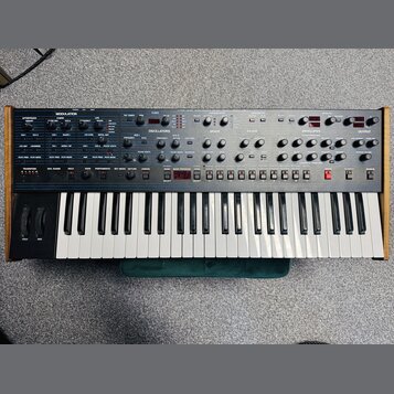 Široký analogový synthesizér s klaviaturaí a dřevěným bočním rámem. Horní panel je šedý s mnoha ovládacími knoflíky a přepínači, rozdělenými do sekcí…