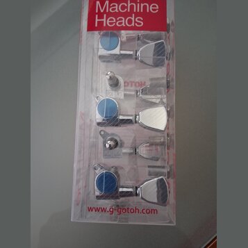 Sada ladících mechanismů v plastovém obalu s červeným nápisem Machine Heads. Mechanizmy jsou chromované s modrými knoflíky a na spodní části obalu je…
