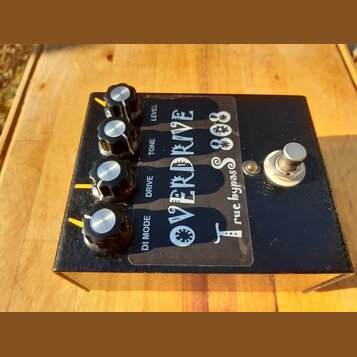 Černý kytarový efekt s nápisem OVERDRIVE TS 808 a doplňkem true bypass. Na horní straně jsou čtyři černé otočné knoflíky s kovovými vršky a popisky L…