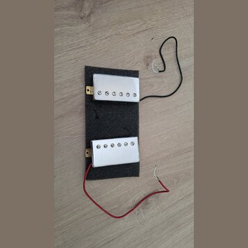 Dvě elektrické kytarové snímače s chromovaným krytem, které jsou připevněny k šedému pěnovému podkladu. Snímače mají pět viditelných šroubů v řadě. Z…
