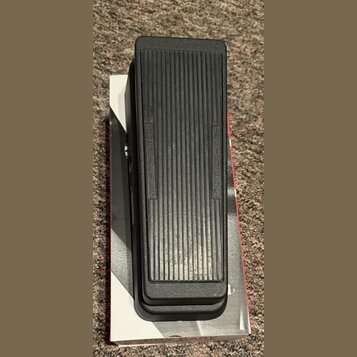 Černý sustain pedál s žebrovaným povrchem, umístěný na bílé krabici. Na pedálu jsou vyrazeny nápisy INSTRUMENT a AMPLIFIER. Povrch plastu vykazuje mí…