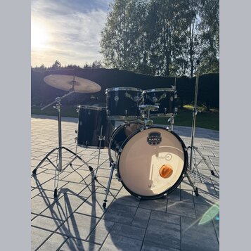 Černá bubnová sada s chromovaným kováním, zahrnující basový buben, dva tom-tomy, floor tom a jeden činel. Na přední bláně basového bubnu je logo MAPE…