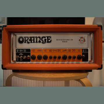 Kytarový zesilovač v oranžovém texturovaném potahu s kovovými úchyty po stranách. Na horním bílém panelu je velký nápis ORANGE a text ROCKERVERB 100…