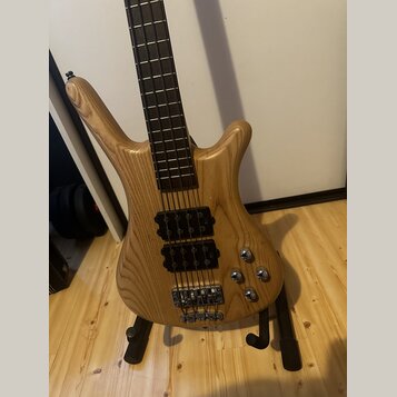 Světle hnědá elektrická basová kytara s přírodným dřevem a viditelnou masivní strukturou zrna. Nástroj má dva černé humbuckery, čtyři ovládací knoflí…