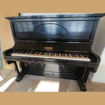 Černý piano (pianino) s otevřenou horní deskou, která obsahuje oválný reliéfní ornament. Nad klávesnicí je umístěno zlaté logo s nápisem "Bechstein".…
