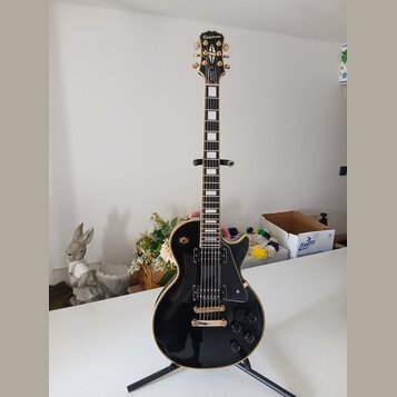 Černá elektrická kytara typu Les Paul s bílými hrankami a zlatým hardwarem. Na hlavě je nápis Epiphone a logo v podobě křídla. Kytara má dva humbucke…
