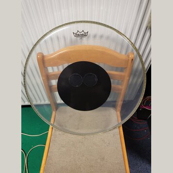 Průhledná bubenová blána s černým kruhem uprostřed a dvěma malými otvory. V horní části je černé logo REMO s nápisem CONTROLLED SOUND a pod ním nečit…