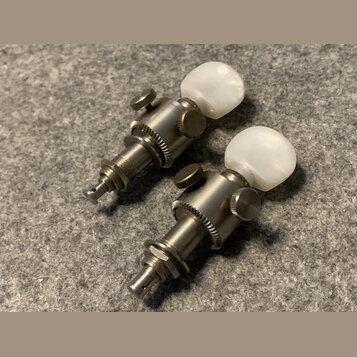 Dvě ladicí mechaniky z kovu s bílými plastovými knoflíky ve tvaru kuliček. Povrch kovových částí vykazuje známky opotřebení a oxidace. Na mechanikách…