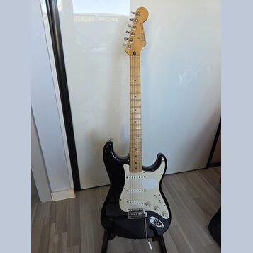 Černá elektrická kytara s bílým pickguardem a hnedým javorovým hridlem. Na hlavě je viditelné logo Fender a nápis Stratocaster. Nástroj vykazuje znám…