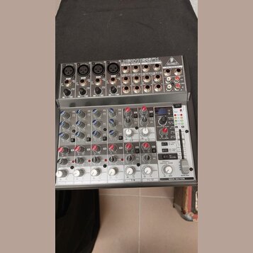 Stříbrný mixážní pult s nápisem BEHRINGER XENYX 1202FX. Zařízení obsahuje několik vstupních kanálů s potenciometry, přepínači a konektory typu XLR a…