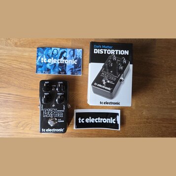 Na fotografii je kytarový efekt Dark Matter od značky tc electronic, jeho originální krabice a manuál. Pedal má černé kovové tělo s bílým nápisem DAR…