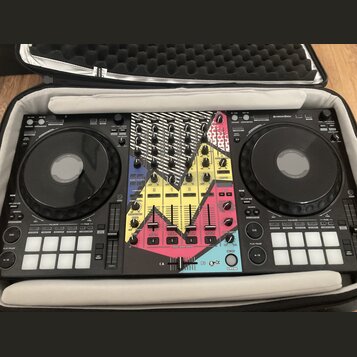 Černý DJ 컨트롤러 umístěný v přepravním kufru. Střední část přístroje je pokryta barevnou polepkou s růžovými, žlutými a modrými geometrickými tvary. V p…