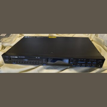 Černý rackový CD přehrávač s nápisem TASCAM CD-500B a označením MP3 WAV. Zařízení má digitální displej, ovládací tlačítka s číslicemi 0–9 a funkční p…