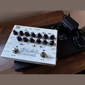 Bílý akustický předzesilovač s nápisem EBS a Stanley Clarke Signature ACOUSTIC PREAMP. Zařízení má několik ovládacích knoflíků, přepínačů a vstupů/vý…