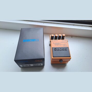 Na fotografii je oranžový kytarový efekt BOSS TURBO Distortion DS-2 v originální černé krabici. Na pedálu jsou viditelné čtyři ovládací knofíky, nápi…