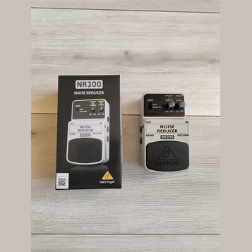 Na fotografii je šedý kytarový pedál Noise Reducer značky behringer a jeho originální kartonová krabice. Na pedálu i na krabici je uveden model NR300…