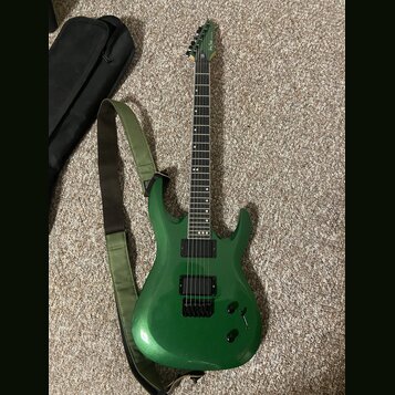 Zelená elektrická kytara s kovovými strunami a dvěma černými humbuckery. Na hlavě je viditelné logo Ibanez. Kytara má zelený popruh a vedle ní leží č…