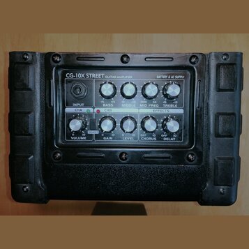 Kytarový zesilovač s černým povrchem a ovládacím panelem. Na panelu je nápis CG-10X STREET GUITAR AMPLIFIER a BATTERY & AC SUPPLY. Ovládání obsahuje…