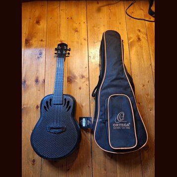 Černá kytara s povrchem imitujícím karbonové vlákno a doprovodný černý pouzdro s oranžovým lemováním. Na pouzdře je bílé logo a nápis ORTEGA GUITARS…