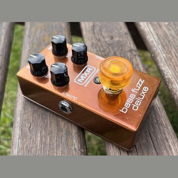 Bronzový kytarový efekt s nápisy MXR, BASS INNOVATIONS a bass fuzz deluxe. Zařízení má čtyři černé plastové knofíky a jeden velký průsvitný oranžový…