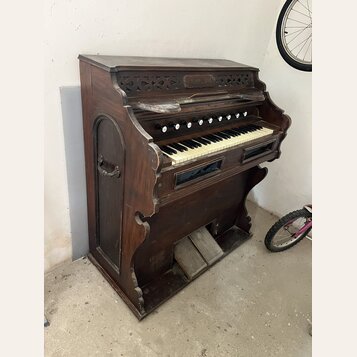 Hnědý dřevěný pomníkový orgán s klaviaturaí. V horní části je vyřezávaný ornament a nápis "SAYER & SON". Povrch dřeva vykazuje známky opotřebení a od…