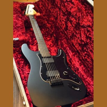 Černá elektrická kytara s matným povrchem v červeném plišovém kufru. Nástroj má dva humbuckery, černý pickguard a světlou hmatnici. Na hlavě kytary j…