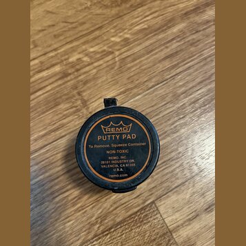 Kulatá černá plastová nádobka s oranžovým potiskem. Na víčku je logo koruny a nápisy REMO, PUTTY PAD, To Remove, Squeeze Container, NON-TOXIC, REMO,…