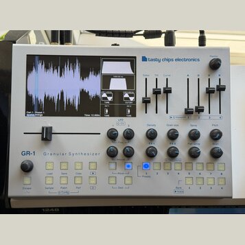 Stříbrný granulární syntetizátor s nápisem GR-1 Granular Synthesizer a logem tasty chips electronics. Zařízení má displej s vizualizací vlnovky, něko…