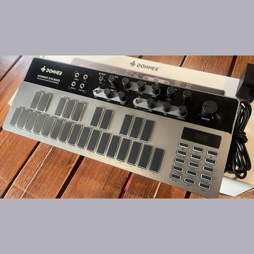 Stříbrný analogový syntetizátor s černým ovládacím panelem. Na horní části je nápis DONNER SYN BASS ANALOG SYNTHESIZER AND SEQUENCER a logo DONNER. P…