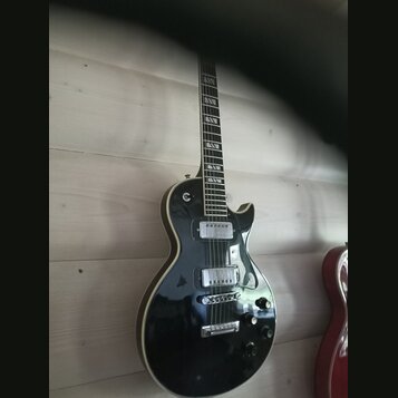 Černá elektrická kytara s tvarem single-cutaway a bílým lemováním kolem korpusu. Nástroj má dva humbuckery, Tune-o-matic můstek a čtyři ovládací knof…