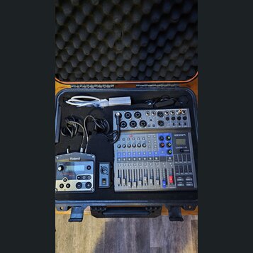 V černém přepravním kufru s pěnovou výplní se nachází digitální mixáž ZOOM LiveTrak L-8, modul Roland TR-505 a bílý napájecí adaptér s kabelem. Na mi…