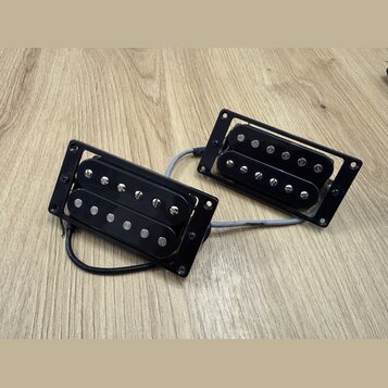 Dvě černé humbuckery s montážními rámečky, položené na dřevěném povrchu. Snímače mají viditelné kovové šroubky a pólové piny. Na fotce nejsou žádné č…