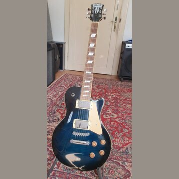 Tmavě modrá elektrická kytara s tvarem singlecut a bílým pickguardem. Na hlavě je viditelné logo Gibson. Kytara má dva humbuckery s kovovými kryty, č…