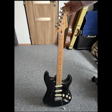 Černá elektrická kytara s bílým pickguardem a javorovým hnedým hmatníkem. Na hlavě je viditelné logo Fender a nápis STRATOCASTER. Kytara má tři sníma…