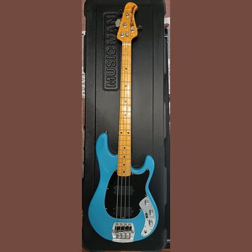 Modrá elektrická basová kytara v černém tvrdém kufru s nápisem MUSIC MAN. Nástroj má světle hnědý hриф z dřeva a černý pickguard. Na hlavě je viditel…