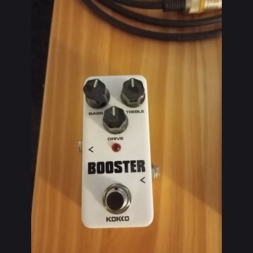 Bílý kytarový efekt s nápisem BOOSTER a značkou KOKKO. Na horní straně jsou tři černé otočné knoflíky s popisky BASS, DRIVE a TREBLE. Uprostřed se na…