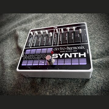Kytarový efekt v bílém šasi s černým horním panelem. Na panelu je nápis MICROSYNTH a logo electro-harmonix s doplňkem MADE IN NYC, USA. Horní část ob…