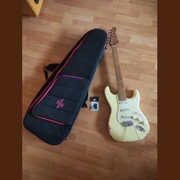 Bílá elektrická kytara s jasným hnedým hřidnicem a bílým pickguardem. Na hlavě je viditelné logo Fender. Vedle kytary leží černý obal s růžovým lemov…