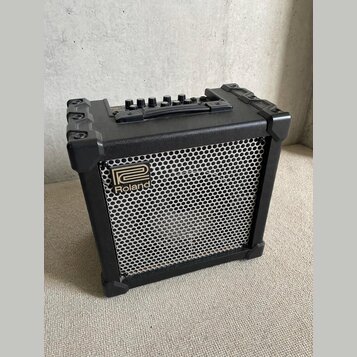 Černý kompaktní kombo zesilovač s kovovou mřížkou reproduktoru. Na přední straně je umístěno logo a nápis Roland. Horní panel obsahuje ovládací knofl…