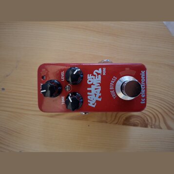 Červený kytarový efekt s nápisy HALL OF FAME 2 MINI a tc electronic. Na horní straně jsou tři černé otočné oválné knoflíky s popisky LEVEL, TONE a ne…