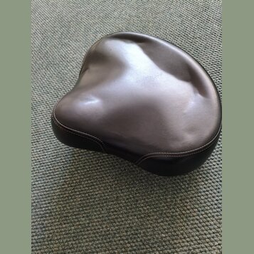 Černý polstrovaný sedák v ergonomickém tvaru. Povrch je z hladkého materiálu s viditelným prošíváním po okraji. Na fotce nejsou žádné viditelné značk…