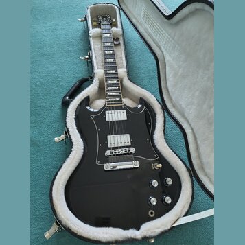 Černá elektrická kytara s dvojstupňovými humbuckery a bílým pickguardem, umístěná v otevřeném černém kufru s bílou vnitřní výstylou. Na hlavě nástroj…
