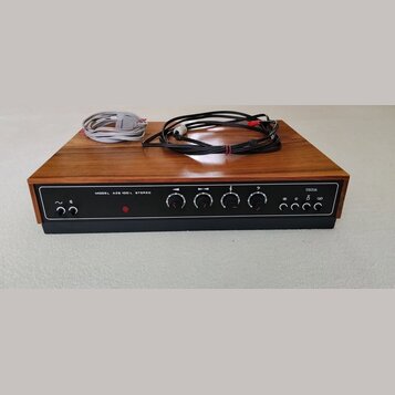 Stereo zesilovač v dřevěném šasi s černým čelním panelem. Na panelu je nápis MODEL AZS 100 L STEREO a logo TESLA. Jsou zde čtyři otočné ovladače, něk…