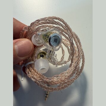 Sluchátka in-ear s průhledným plastovým tělem a měděně zbarveným pleteným kabelem. Na vnitřní straně jednoho sluchátka je viditelné černé kulaté logo…