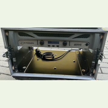 Šedý rackový zesilovač s nápisy crown a 4002 XTi SERIES. Zařízení má dva otočné ovladače, displej a několik tlačítek. Je umístěno v transportním kufr…