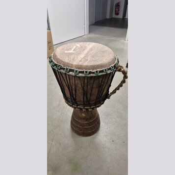 Djembe s tělem z tmavého dřeva a koženou blánou. Horní okraj je opleten zeleným šňůrkem a nástroj je napnutý černými provazníky. Na pravé straně je p…