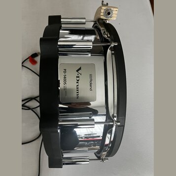 Elektronický snare s chromovaným kovem a černými plastovými prvky. Na boku je vidět stříbrný štítek s nápisy Roland, V-Drums a PD-140DS V-PAD DIGITAL…