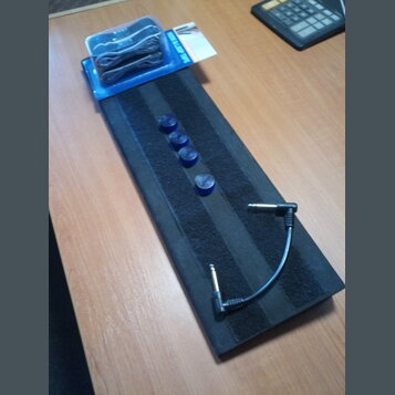 Na černém podkladu leží krátký černý audio kabel s 6,3mm jack konektory na obou koncích. Vedle něj jsou čtyři modré plastové krytky a v horní části j…
