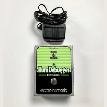 Kytarový pedál značky electro-harmonix s názvem Hum Debugger a přívostkem Hum Eliminator. Zařízení má stříbrný kovový šasi, zelený vršek s vodorovným…